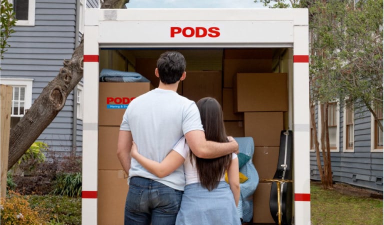 A couple is standing with their arms around each other, looking at their PODS moving container that they've just finished loading with moving boxes