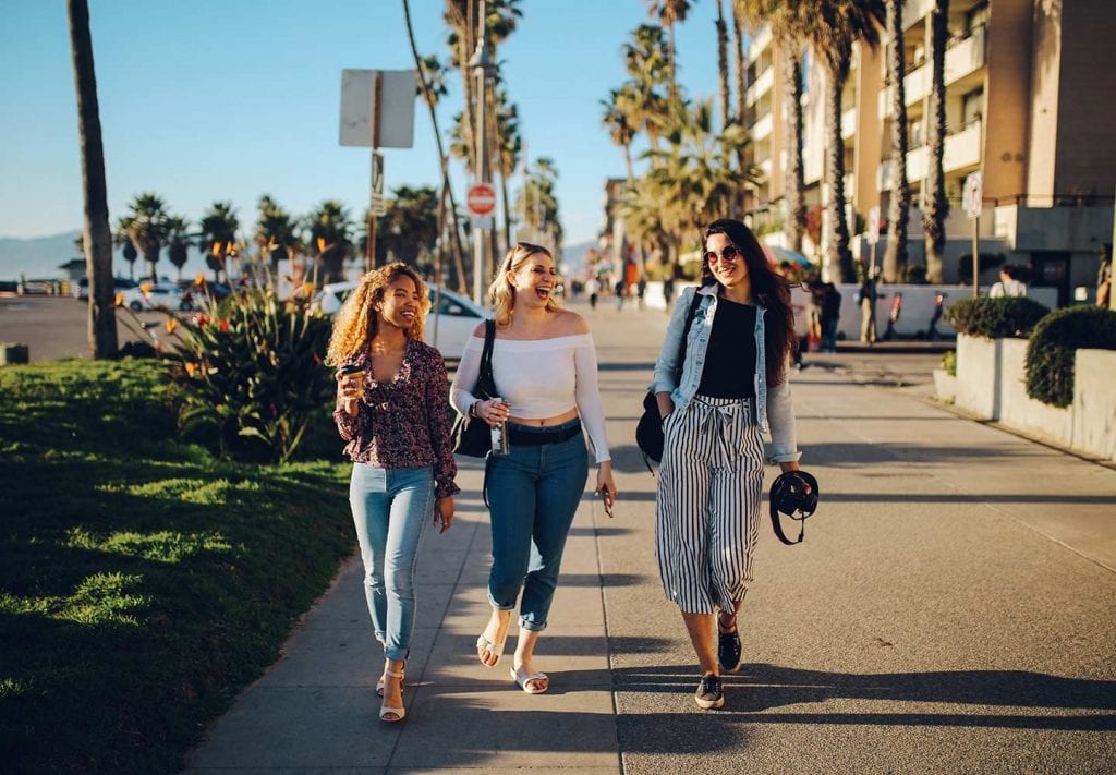 A trio of young women walk along the sidewalk at a beach. They are laughing. 