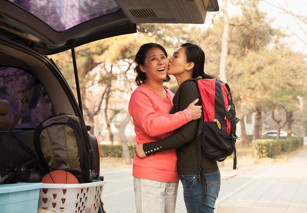 A daughter kisses her mother as they move to Los Angeles. The daughter is wearing a red backpack and the mom stands next to a car with an open trunk. 