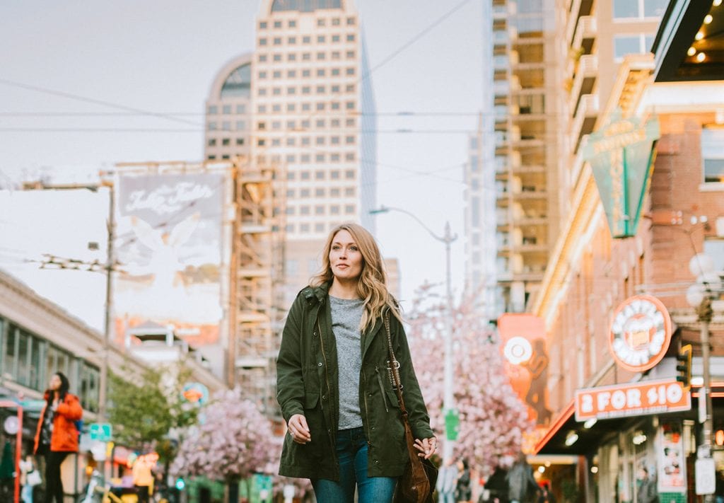 A young woman walking through the city streets of Seattle on a clear day. She’s wearing a fall jacket and there’s activity on the sidewalks behind her.