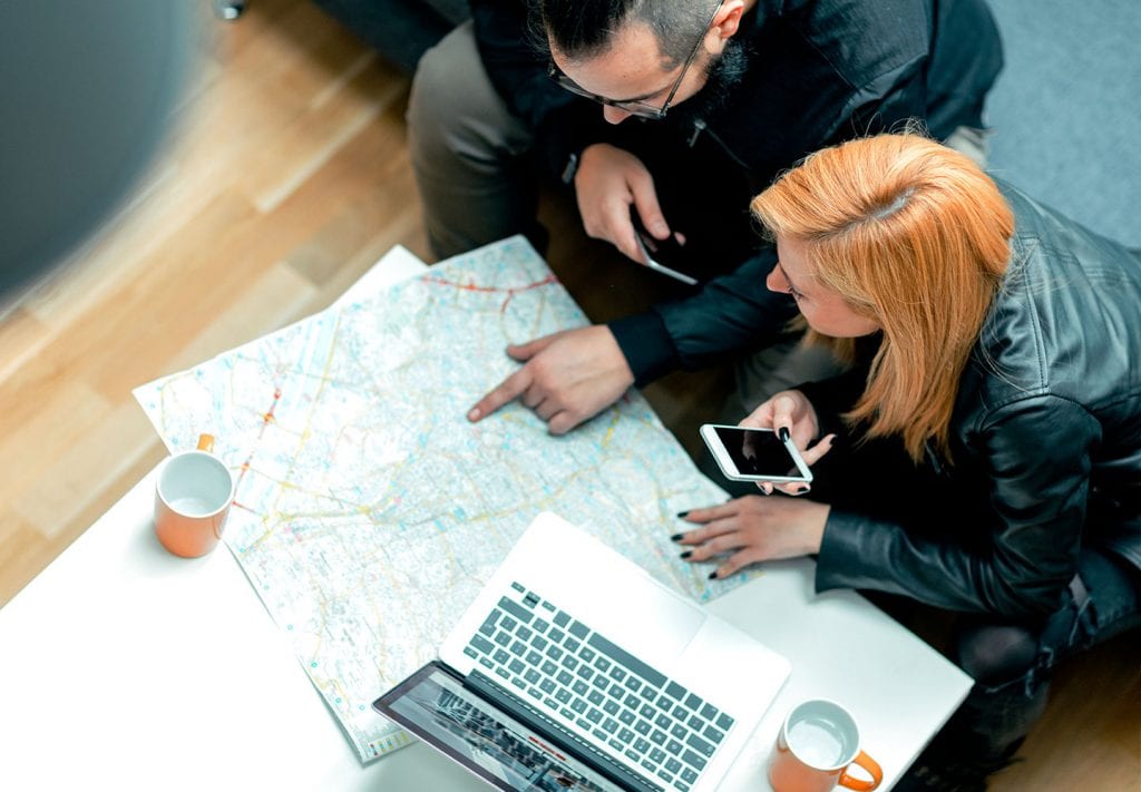 A couple is sitting at a table looking over a map of the Seattle area. They’re each holding their mobile phones and there’s a laptop open on the table. They’re trying to find an affordable Seattle neighborhood to move to.