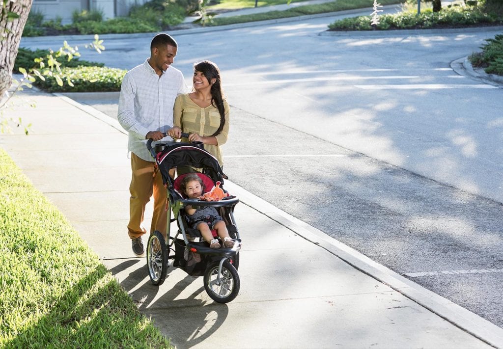 A couple walks their baby in a stroller down a residential Miami sidewalk. They look at each other smiling. 