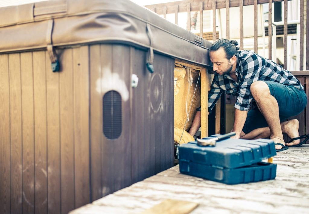 A man in blue shorts and a checked flannel kneels down to clean out the bottom of his hot tub, 