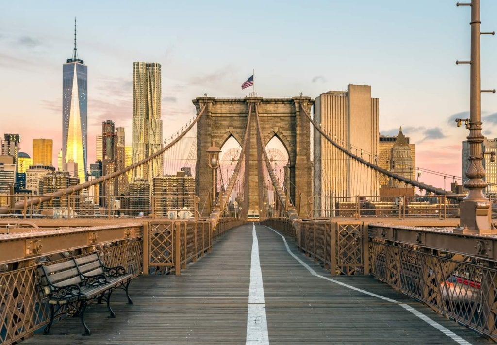 A peaceful view of an empty Brooklyn Bridge with the sun reflecting off of the NYC buildings behind it.