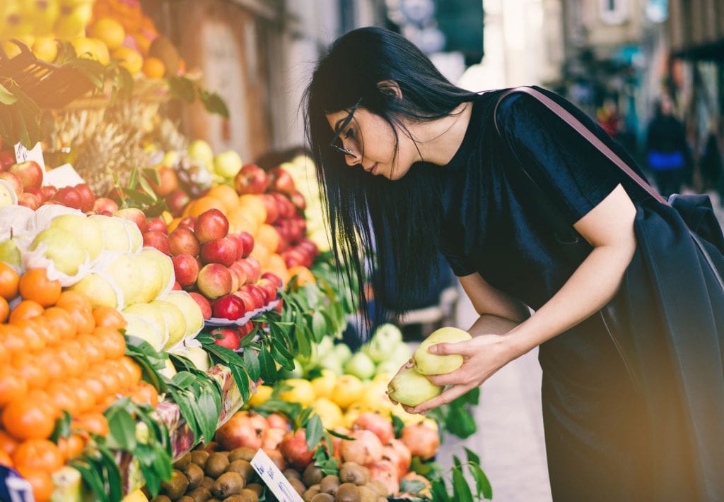 A woman is picking out fresh produce from a local farmer's market stand in NYC. She has two green pears in her hands and is looking at the other colorful fruit options in front of her.