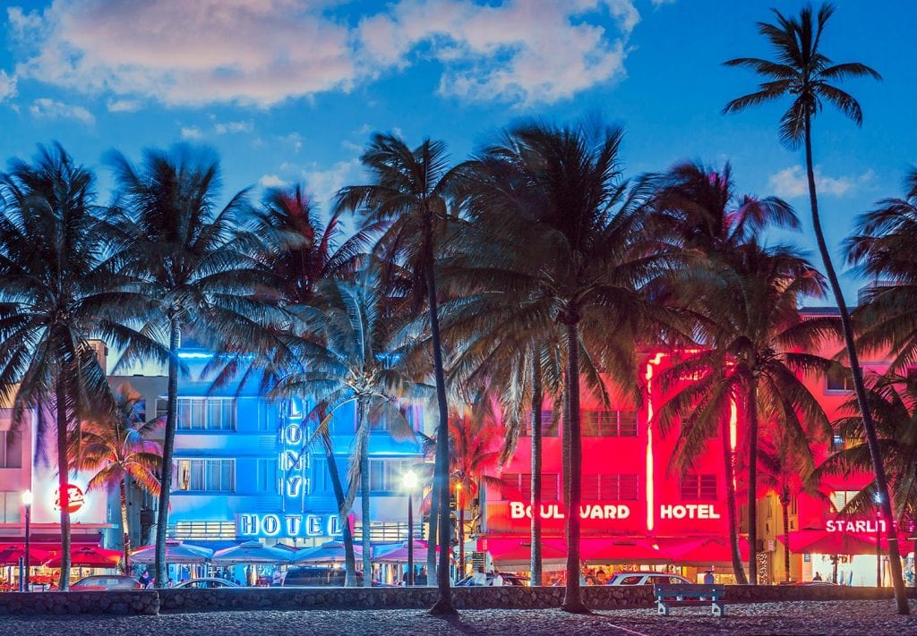 Hotels in South Beach, Miami, at dusk, seen through a line of palm trees from the beach. The hotels are lit up with brightly colored signs.