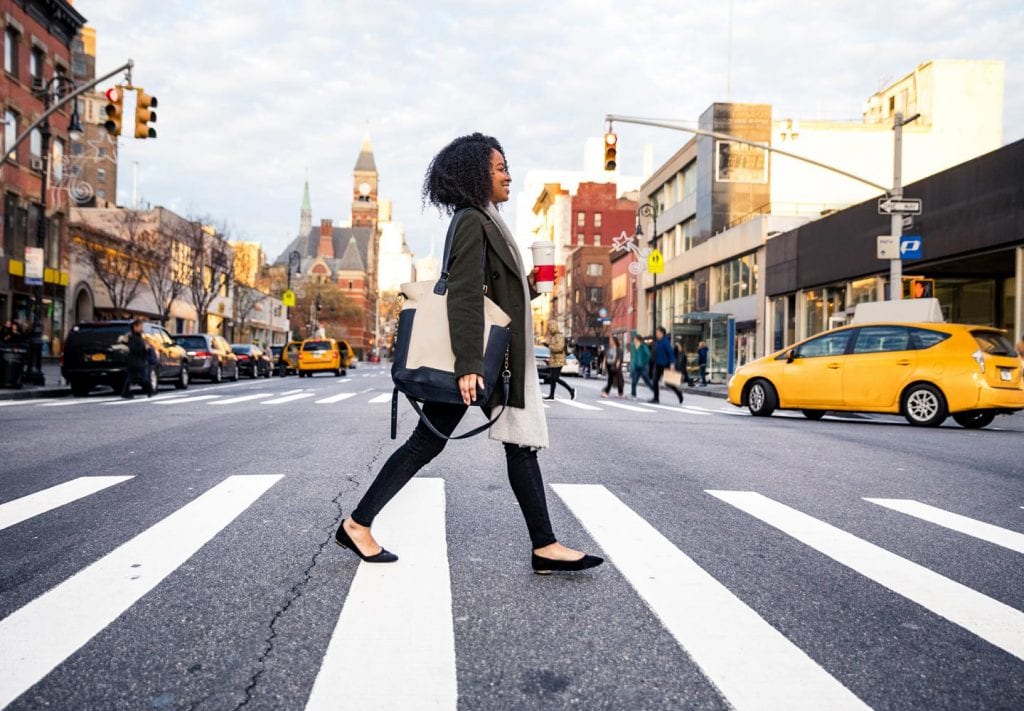 A woman is walking across a crosswalk on a street in New York. She’s carrying a bag and has a big smile on her face. The city is buzzing with activity behind her.