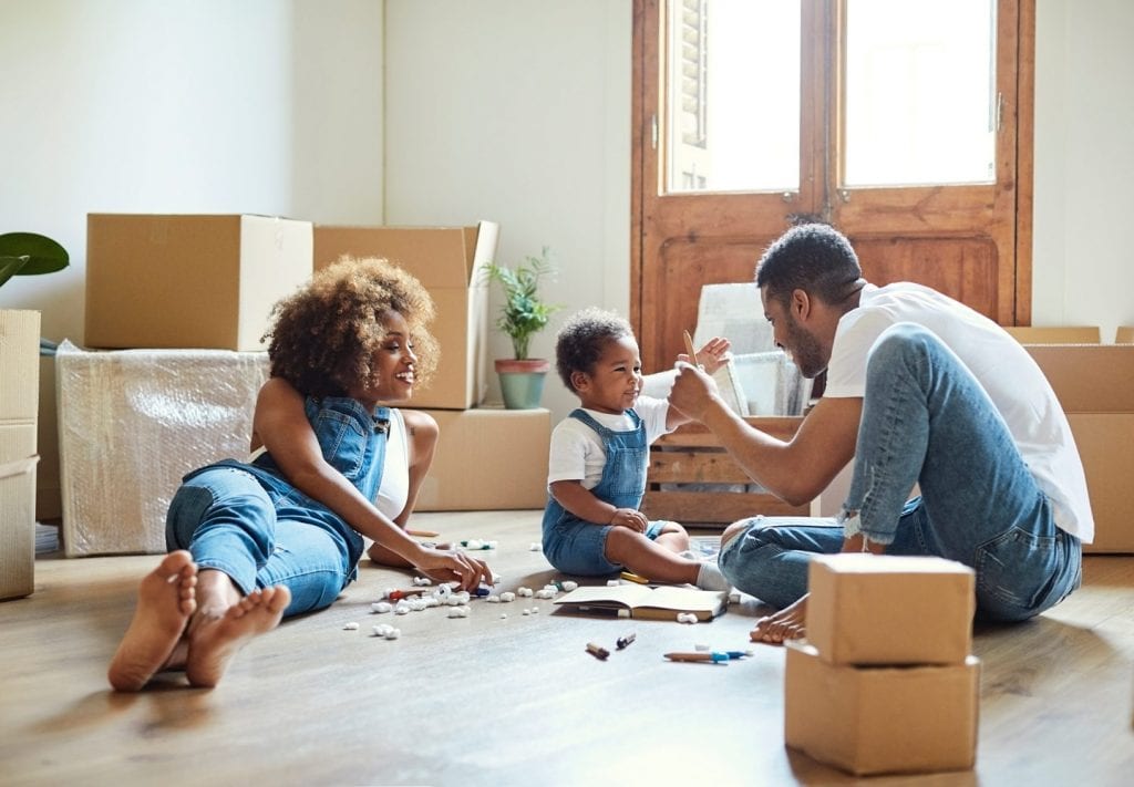 A family is taking a break from unpacking in their new Brooklyn home. The mother, father, and toddler son are reading a book on the floor in the midst of partially unpacked boxes.