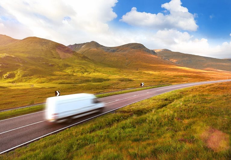 A truck is moving furniture long distance on a deserted two-lane highway in an area with just hills and grass. The truck is in motion, so it’s blurry.