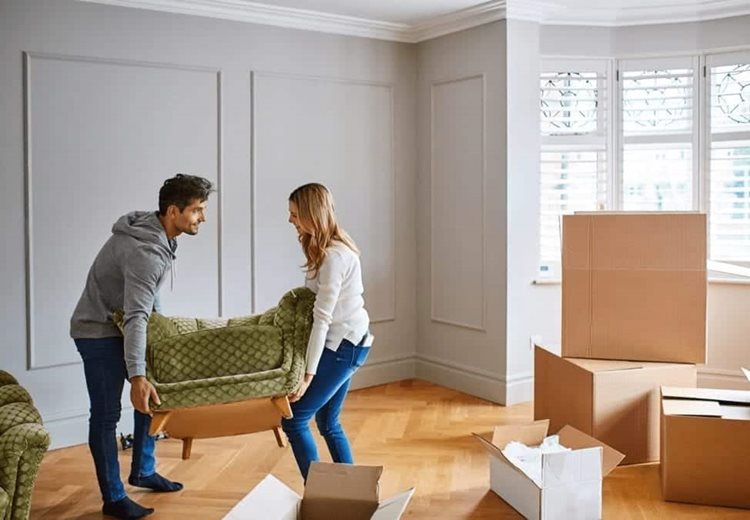 A man and woman are lifting an antique chair. There is a matching sofa to the left of them and several moving boxes to the right of them.