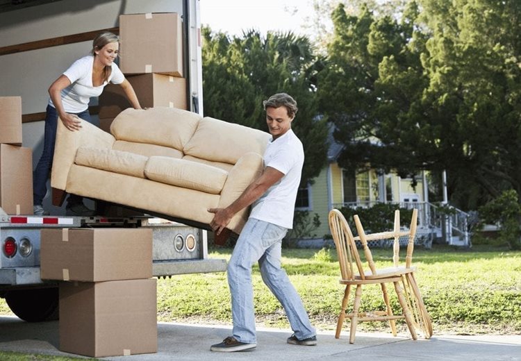 A man and woman are unloading a loveseat from the back of a moving truck after a long-distance move. The man is outside of the truck, and the woman is still in the truck. There are boxes and chairs around them.