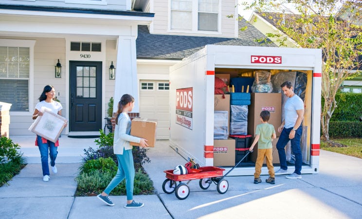 A family of four is carrying things out of their home to load into a PODS portable moving container in their driveway.