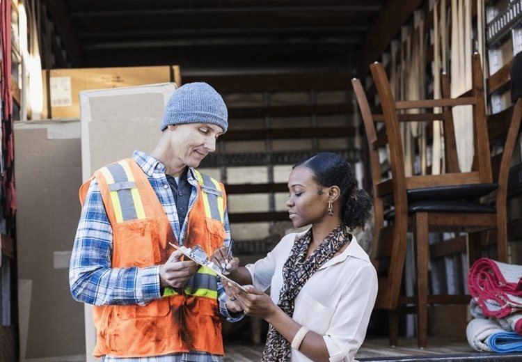 A man with a moving company is getting a woman to sign a bill of lading in front of a moving truck that still has some furniture and boxes on it.