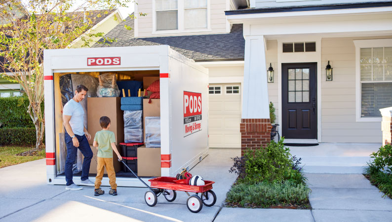 A father and son are talking beside their loaded PODS portable moving container, in the driveway of their home. The boy has a little red wagon.