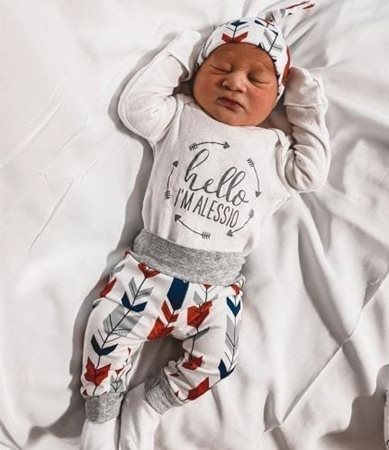 An adorable newborn baby boy is decked out in a cute outfit with decorative arrows on the pants, shirt, and hat. His shirt reads, “Hello I’m Alessio.”