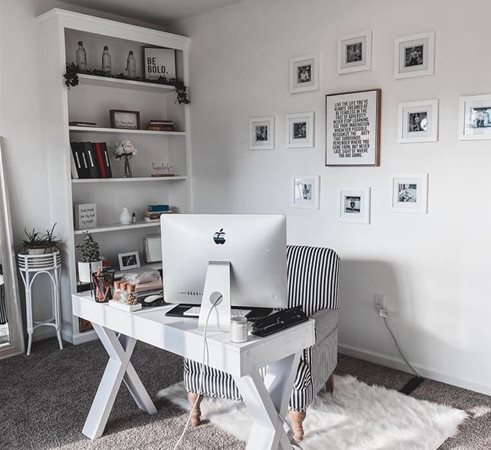 A home office is decorated in a black and white color theme. There’s a chic striped chair at the desk with a furry white rug beneath it. The wall behind the desk features a framed inspirational quote with smaller framed images around it.