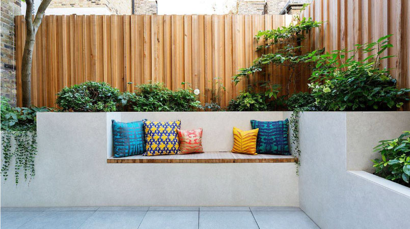 A fenced in patio with tile flooring and bench seating built into the concrete walls. The seat has inlaid wood and several colorful throw pillows. There are lush plants growing all around.