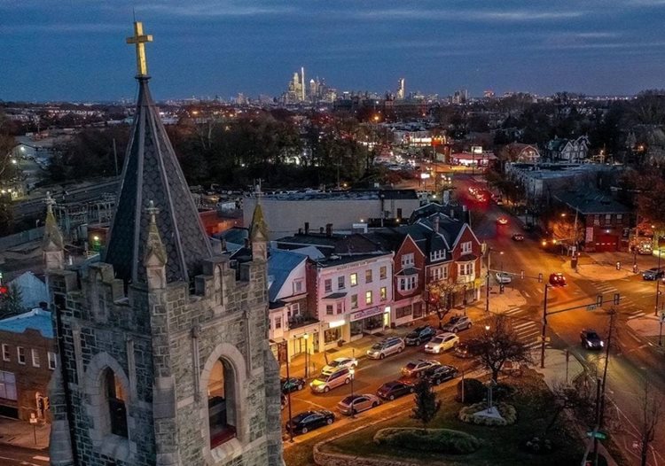 Aerial view of the Philadelphia neighborhood of Overbrook in the evening. In the foreground, the bell tower of a church is visible, surrounded by lower-lying buildings, both residential and commercial. 