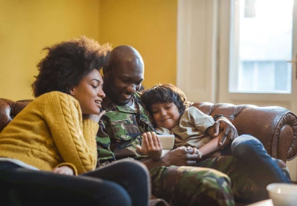 A young military family relaxes on a brown couch while scrolling through a smartphone. 