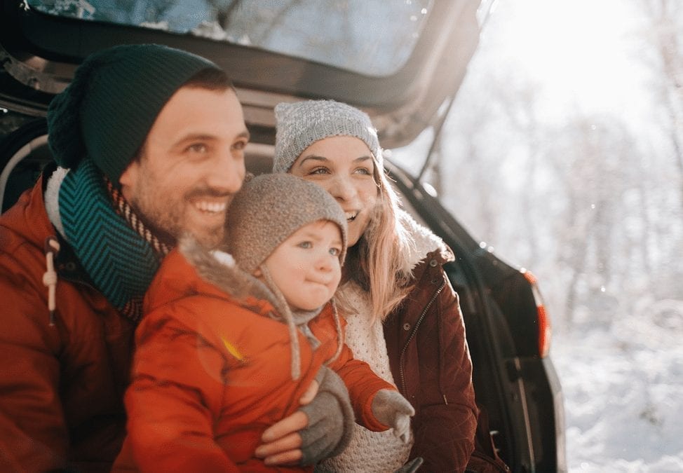 A couple and their toddler are wearing winter clothes and sitting on the back of their car with the hatchback open. They're smiling and looking out at the winter scenery.