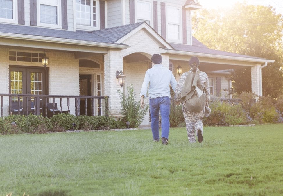 A woman dressed in military fatigues and her husband walk toward a large home hand in hand. 