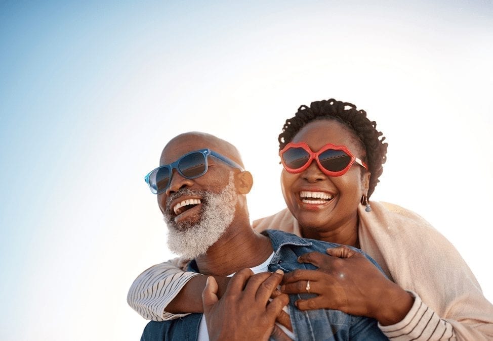 A mature couple is embracing and smiling in warm, sunny weather. They're both wearing sunglasses. 