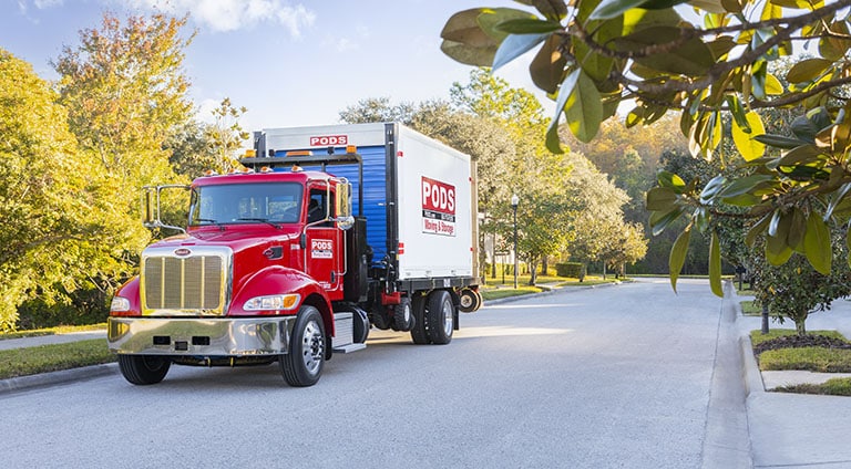 A truck drives a PODS container down a street, ready to move.