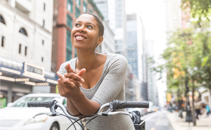 A happy young woman is leaning on her bicycle handles while waiting at a stop light in Chicago. She is smiling and looking out, thinking about something pleasant.