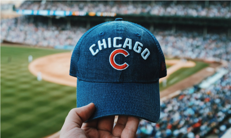 The hand of a Cubs fan, holding their “Chicago C” baseball hat up for a picture in front of a packed Wrigley Stadium.