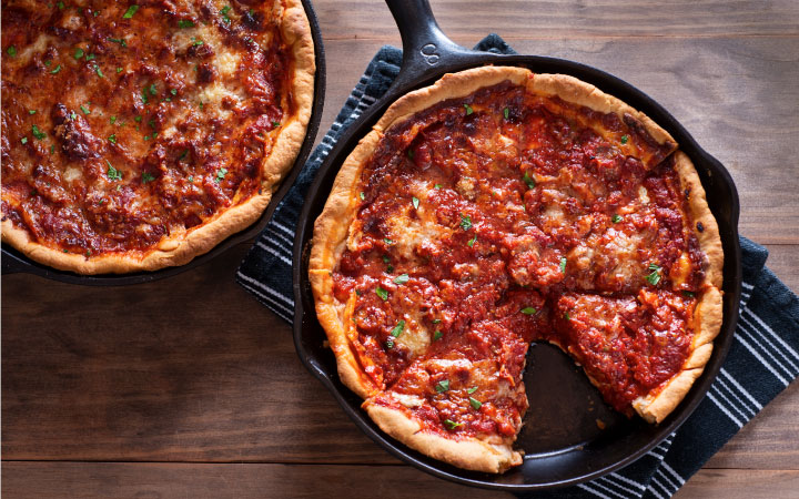 Two pans of Chicago deep-dish pizza are cooling on a wooden table. A slice has been taken from one of the pies.
