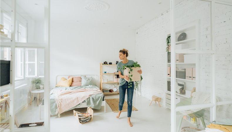 A woman is carrying a square planter with a houseplant in it through her bedroom. She is looking around the white-colored room to decide which pieces of furniture to keep and which ones to replace with modern versions.