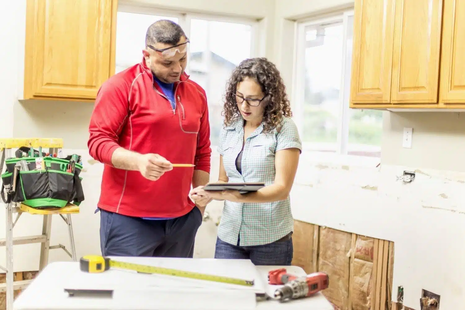A woman reviews her kitchen remodeling plan with the remodeling contractor she hired. They’re standing at the kitchen island, which is covered in paper plans and tools. The kitchen is partially completed.