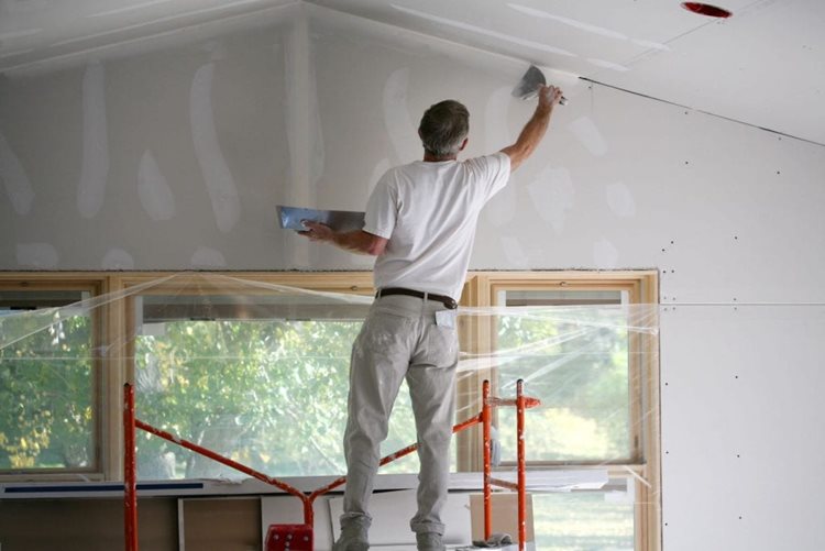 A remodeling contractor is standing on scaffolding, using putty to fill in the cracks between a freshly constructed wall and the ceiling. Plastic sheeting has been secured over windows to protect them.