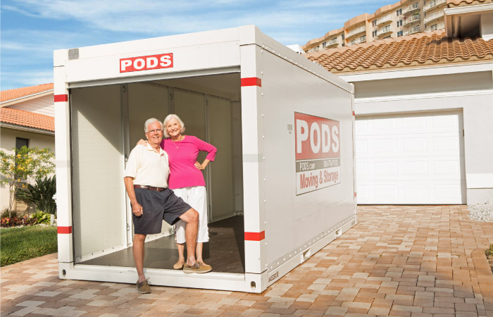 A senior couple is standing together in the front of an empty, open PODS storage container in their driveway.