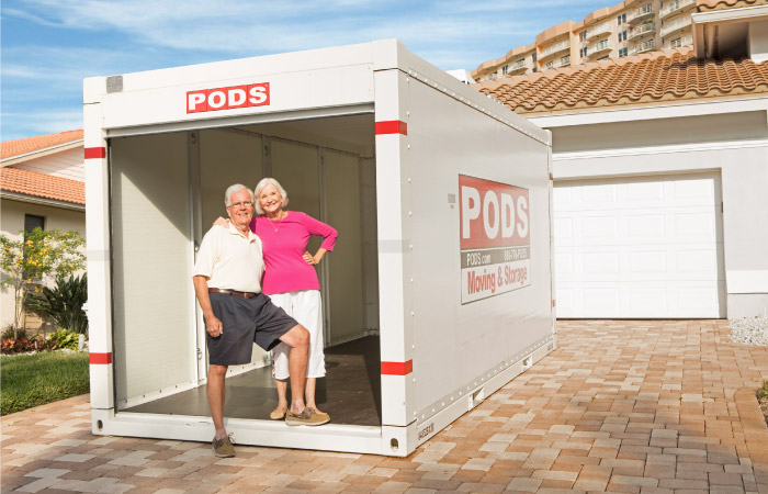  A senior couple stands together in the front of their PODS storage container. The container is positioned in their driveway and the sliding door is open showing that it’s empty inside.