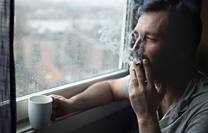 A mustached man is holding a cup of coffee and smoking a cigarette in his apartment. He's looking out the closed window at a gloomy, rainy day. 