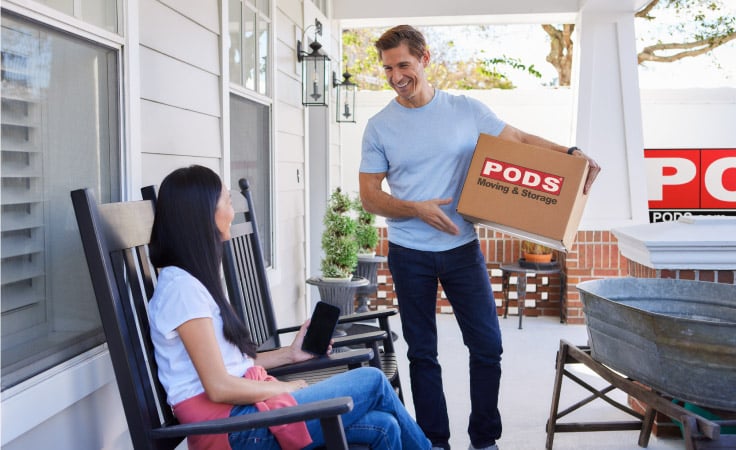 A mature couple smiles while talking on their front porch. The man is holding a moving box labeled “PODS Moving & Storage” and there's a PODS portable moving container in their driveway.