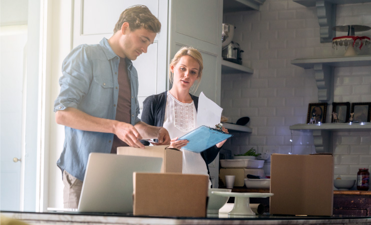 A young couple making an inventory list of the items they plan to sell at their upcoming estate sale. The woman is holding a clipboard with paper and the man is putting a label on a cardboard box. There are two other boxes and an open laptop on the table in front of them.