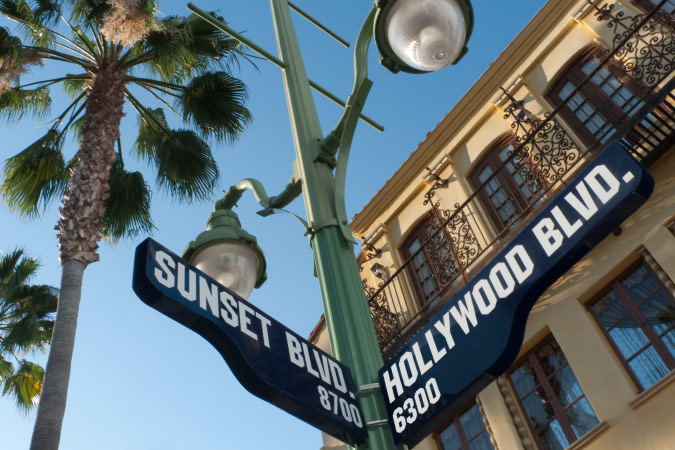View of a pair of street signs at the intersection of Sunset and Hollywood Boulevards in Los Angeles, California, featuring a Spanish-style building and palm trees in the background.