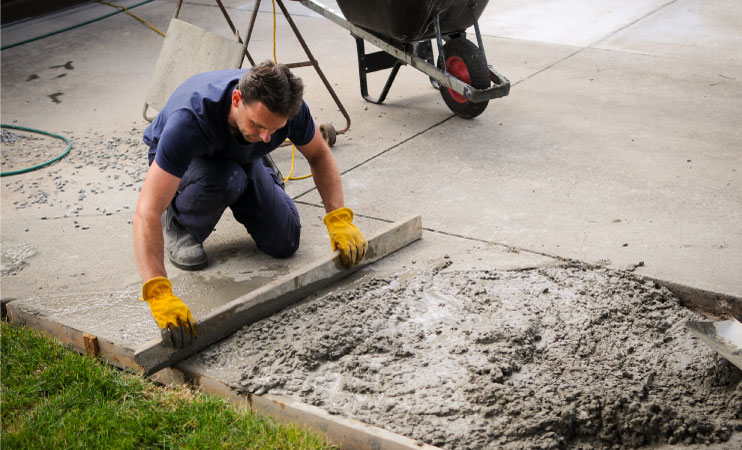 A man is working on a newly poured section of driveway. He’s wearing yellow work gloves and using a 2” by 4” board to smooth out the freshly poured concrete.