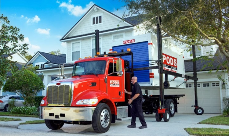 A PODS truck driver is standing beside the cab of his truck. He’s in the process of using PODZILLA to lift a PODS portable moving container off the bed of his truck and then onto the residential driveway.