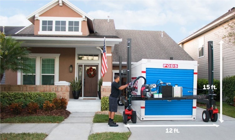A PODS truck driver is using PODZILLA to move a PODS container into place in a residential driveway. There are white graphic lines added to the image showing that one will need a space that is 15 feet tall and 12 feet wide to have a PODS container delivered to their driveway.