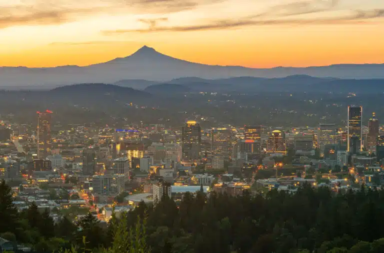 Distant view of the city of Portland, Oregon, during sunset with Mount Hood in the distance and lush, green trees in the foreground.