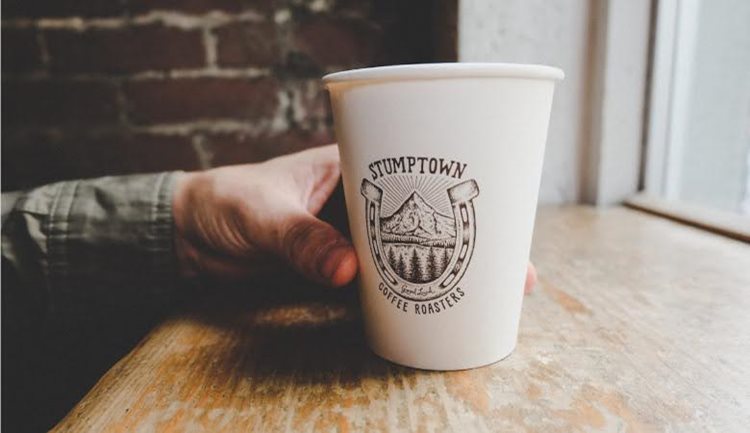 A man’s hand is holding a paper cup of coffee with a decorative stamp on it. The stamp has an illustrated image of Mount Hood, with water and pine trees in the foreground and a large horseshoe surrounding it. The words on the cup read, “Stumptown Coffee Roasters.”