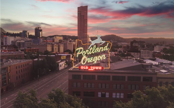 A drone shot of the illuminated “Portland Oregon Old Town” sign in Portland and the surrounding city buildings at dusk. There are wisps of pink and blue clouds in the sky and the leaves on the trees are blowing in the wind.
