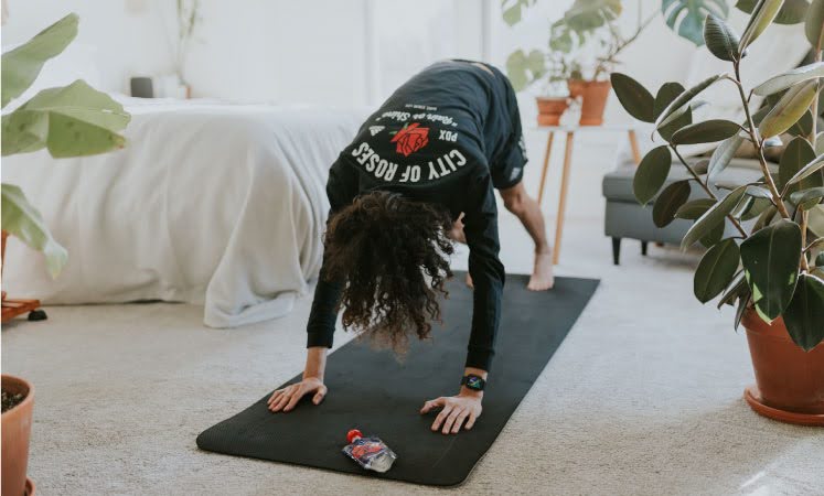 An adult with curly, brown hair is doing yoga in their living room in Portland, Oregon. They’re wearing black shorts and a long sleeve shirt with a picture of a rose and text that reads, “City of Roses,” and “PDX” on it.