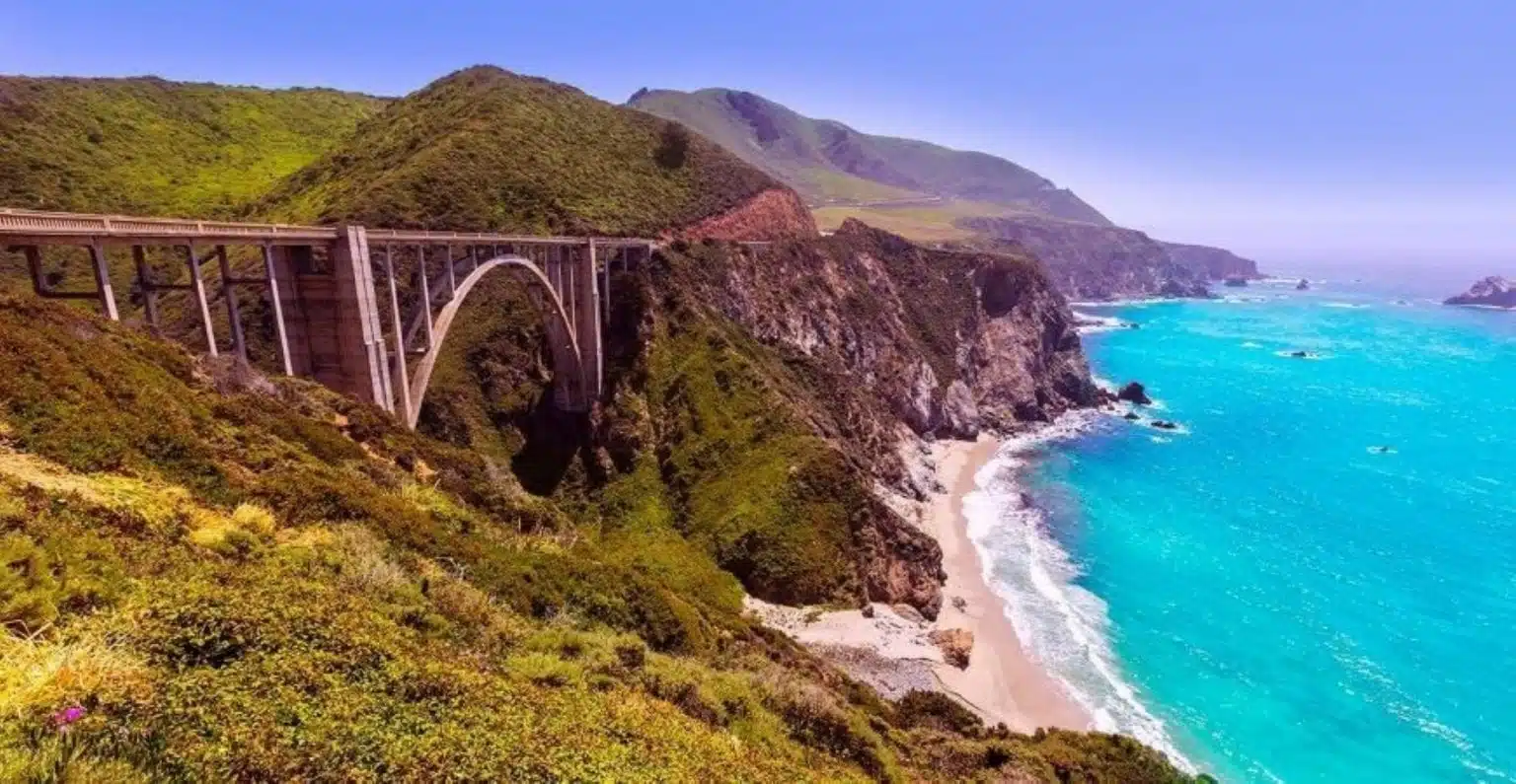 View of a railroad bridge in Big Sur, California, with forested mountains on the left and blue ocean on the right.