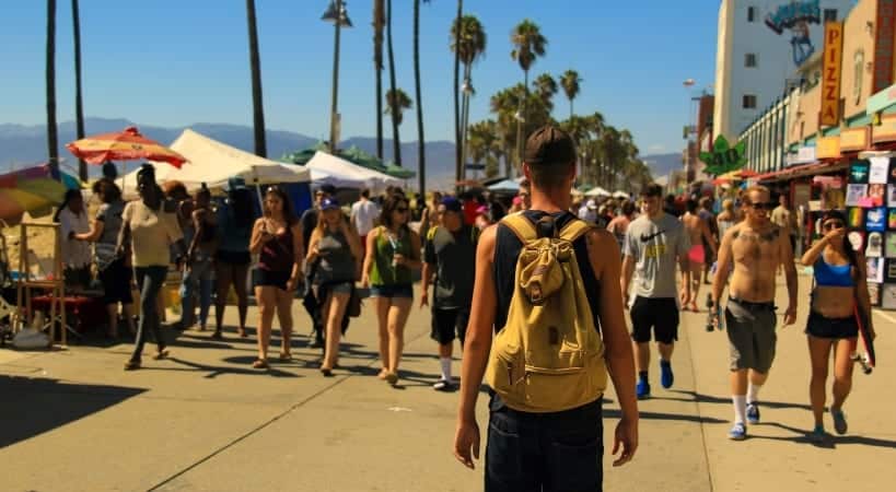 A man is wearing a backpack and standing in the middle of the boardwalk in Venice, California, looking ahead at the crowds of people strolling along the beachfront promenade.