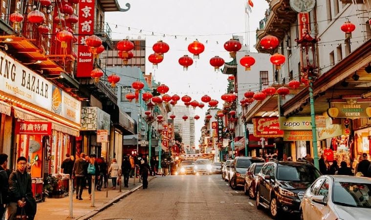 The view down a street in San Francisco’s Chinatown. Red paper lanterns crisscross above the cars, hung from buildings on one side of the street to buildings on the other. Dozens of people stroll along the sidewalk past storefronts. 