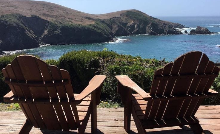 A pair of adirondack chairs are set up beside each other on a wooden deck overlooking the California coast. Waves crash into rocky bluffs, and the water is dark blue. 